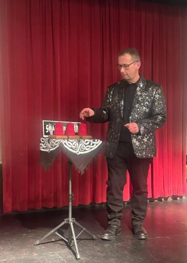 Magician in a black sequined jacket performing on stage with a table and props.